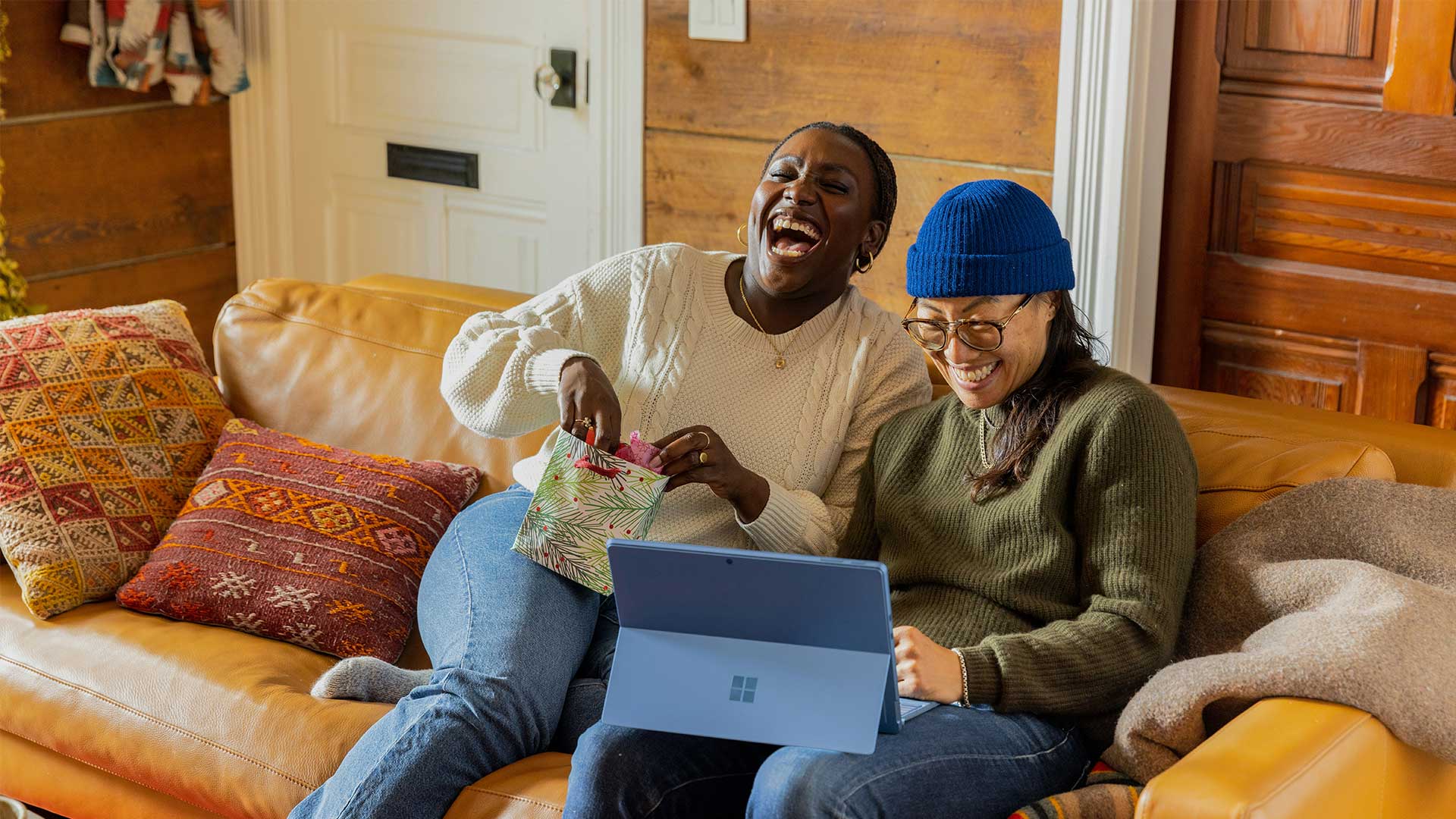 How to live a joyful life: An image of two friends sitting on a yellow sofa. One friend is holding a small gift bag and laughing. The other is smiling at their computer screen.