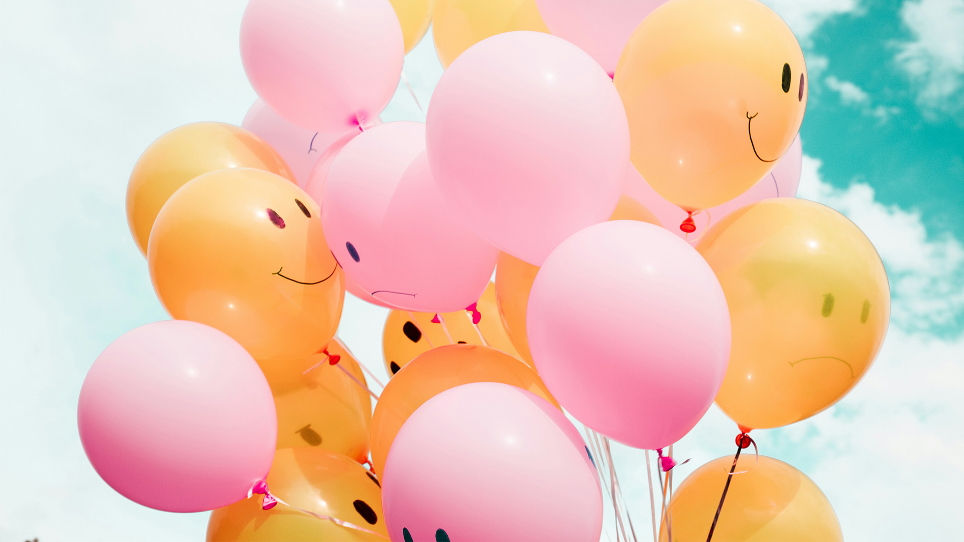 Is it possible to find joy (or at least peace) during difficult times?: An image of pastel pink and orange balloons in front of a cloudy sky. Some of balloons have smiley faces drawn on them, and some have sad faces drawn on them.