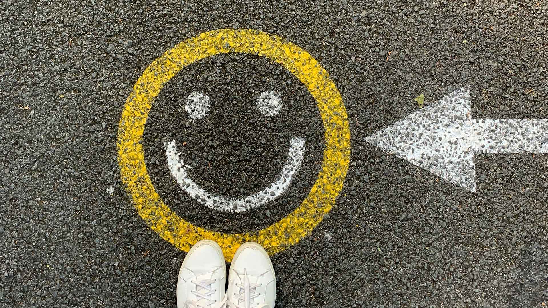 How to live joyfully: An image of a smiley face graffitied onto tarmac. There is a graffiti arrow pointing towards it, with someone's white trainers in the edge of the frame.